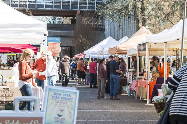People browsing and shopping at an outdoor farmers market with white tents and artisan food stalls.