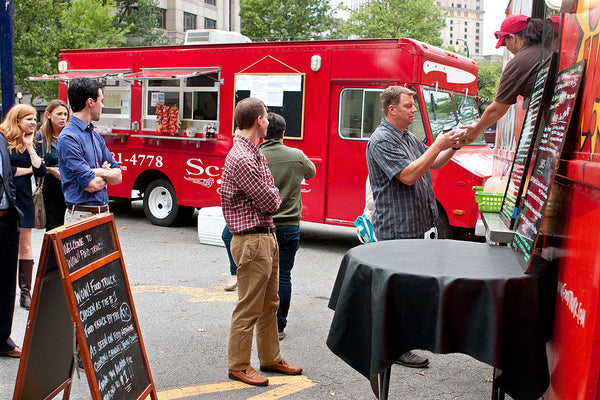 Customers lining up at a red food truck parked on the street during a food festival.