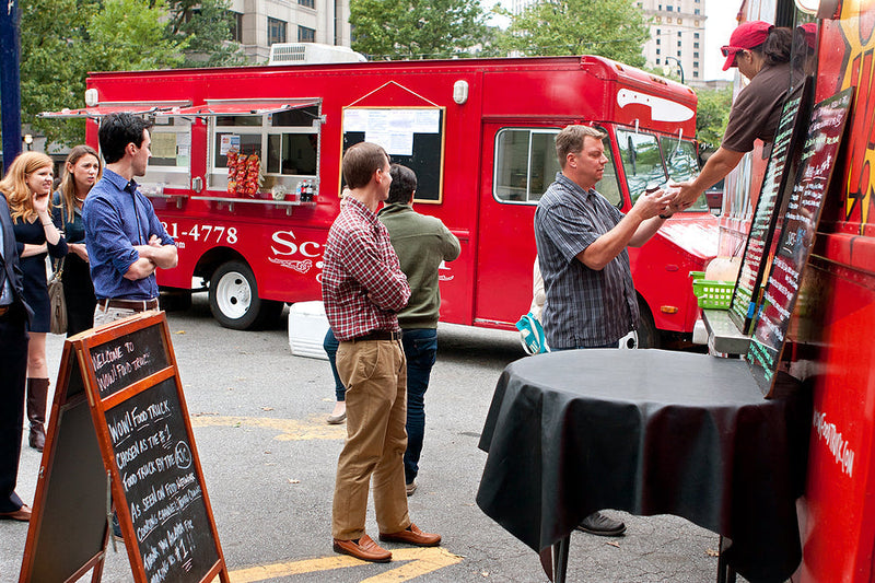 Customers lining up at a red food truck parked on the street during a food festival.