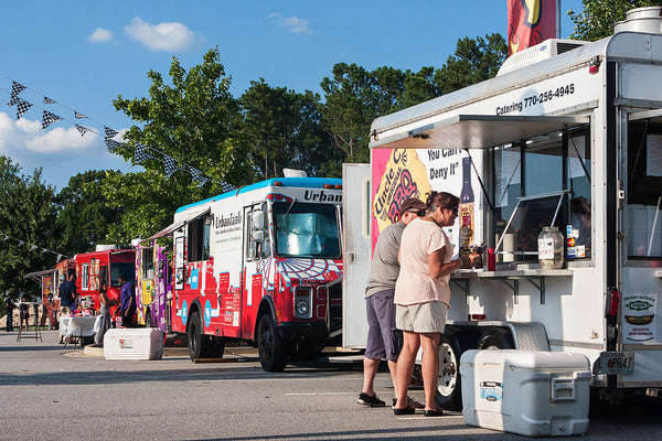 A row of colorful food trucks serving customers outdoors under a sunny blue sky — showcasing vibrant food truck activity during a fall festival.