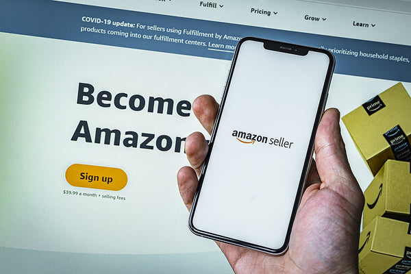 Close-up of a hand holding a smartphone showing the Amazon Seller app screen, with the Amazon Seller sign-up page in the background.